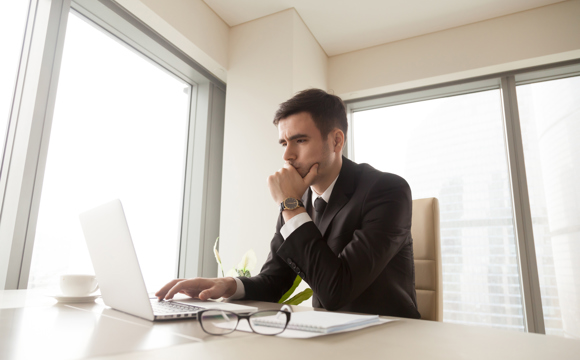 A man in a corner office carefully scrutinizing his laptop