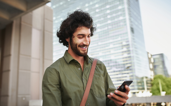 A man in a large city smiling as he uses his phone to check his financial accounts while standing on the street.
