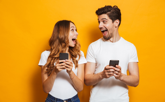 A young man and young woman standing side by side with comically excited smiles as they use their phones.