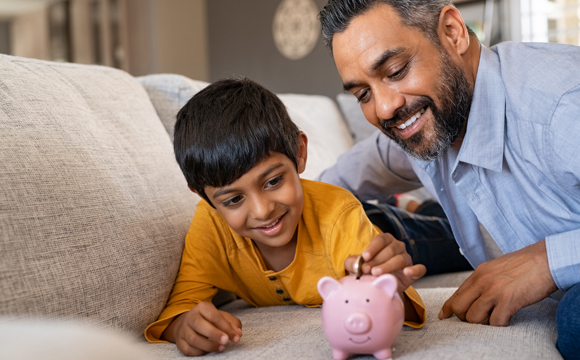 A smiling father and son putting coins into a pink piggy bank together.